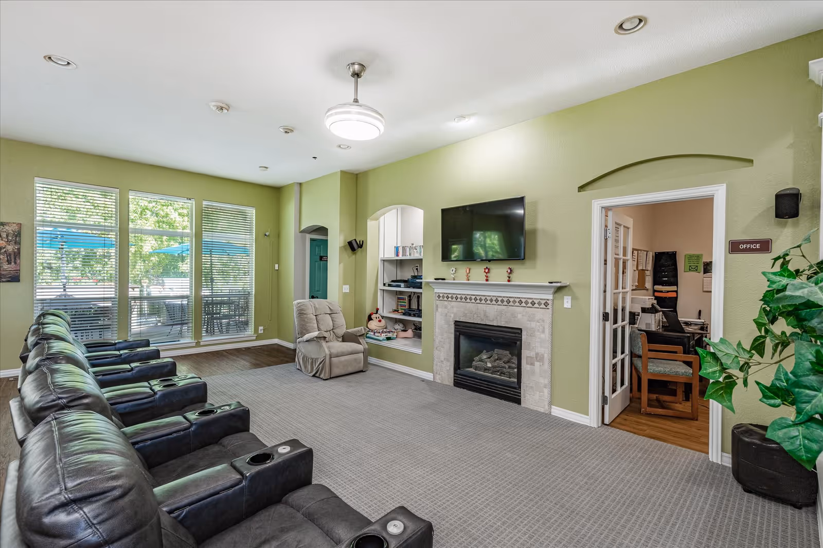A senior living facility common area with a row of black leather recliners facing a fireplace with a mounted flat-screen TV above it. There is a single beige recliner near the fireplace, a built-in shelf with books and decorations, and a door labeled 'Office' leading to a room with office furniture. Large windows with blinds show an outdoor patio with blue umbrellas. The walls are painted light green and the floor is carpeted.