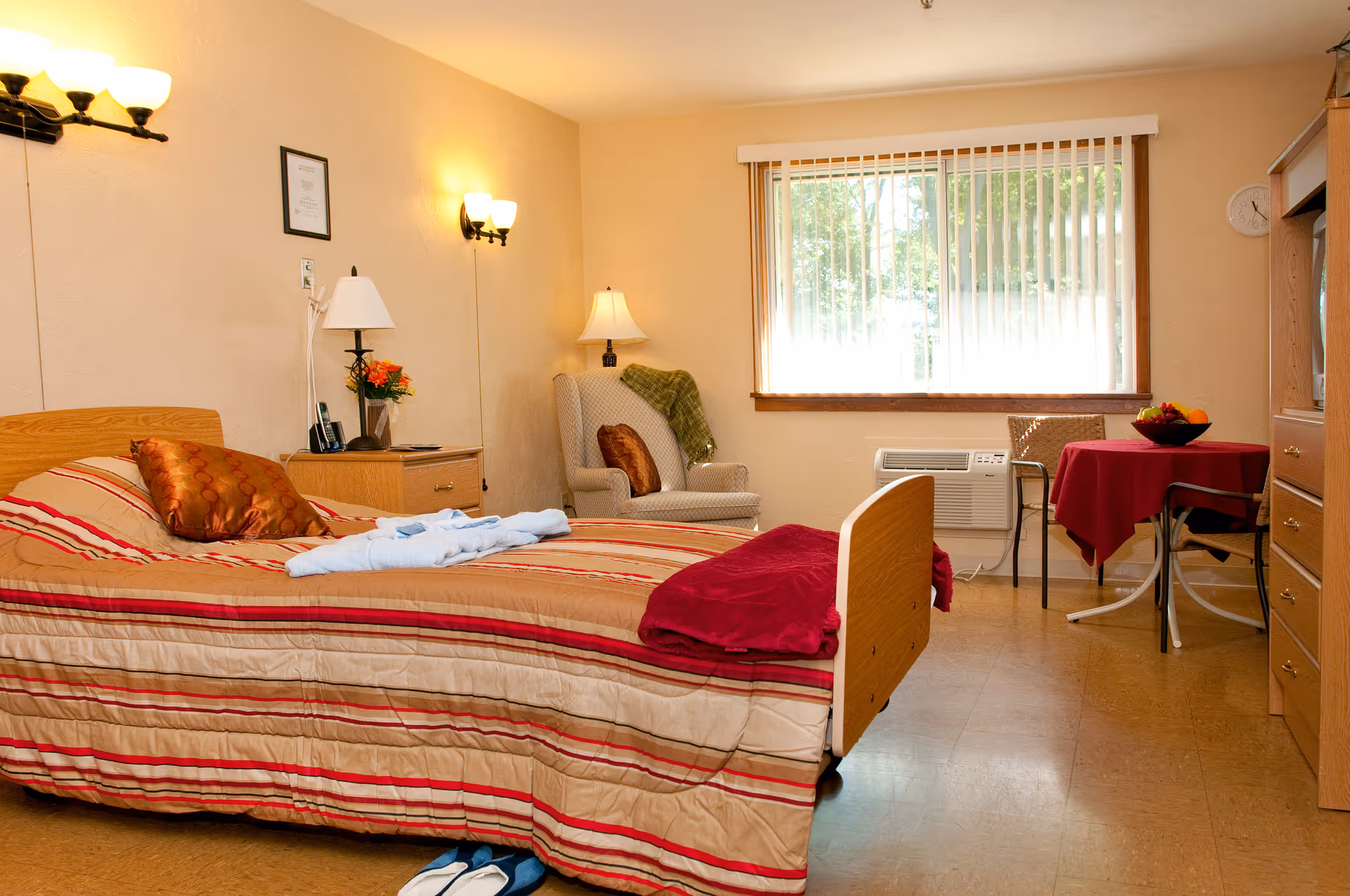 A cozy bedroom in a senior living facility with a single bed covered in a striped beige and red bedspread, a brown pillow, and a folded red blanket at the foot. Next to the bed is a wooden nightstand with a lamp, a phone, and a small flower arrangement. An armchair with a brown pillow and a green throw is positioned near a large window with vertical blinds. A small round table covered with a red tablecloth and two chairs is placed near the window, with a bowl of fruit on the table. The room has warm lighting from wall-mounted lamps and a floor lamp, and a wooden dresser is visible on the right side.