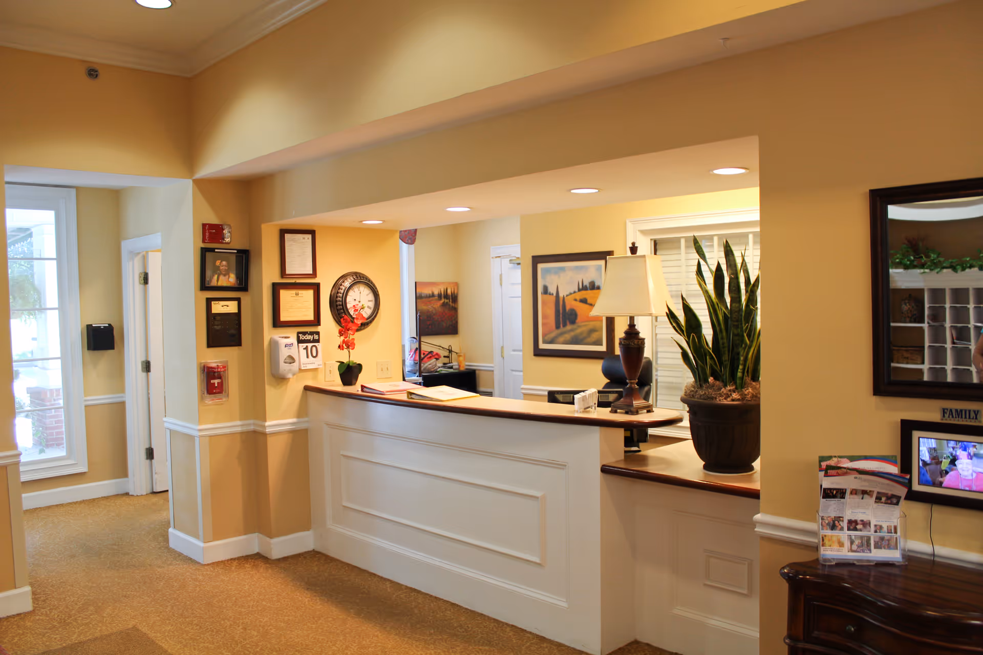 Reception desk and lobby area with a lamp, potted plant, framed art and brochures in a warm-colored interior.
