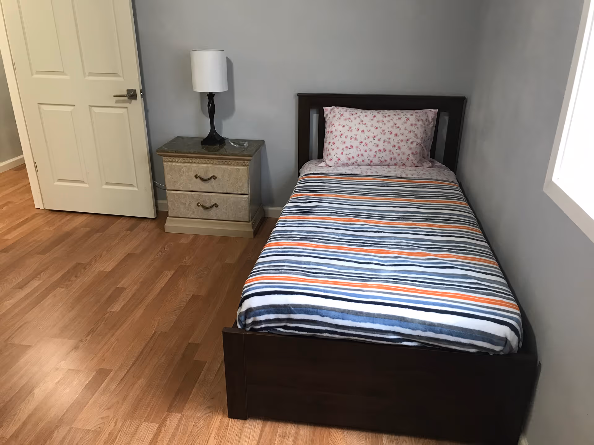 A simple bedroom with a single bed featuring a striped blanket and floral pillow, a nightstand with two drawers, a lamp, and a closed door. The room has wooden flooring and light gray walls.