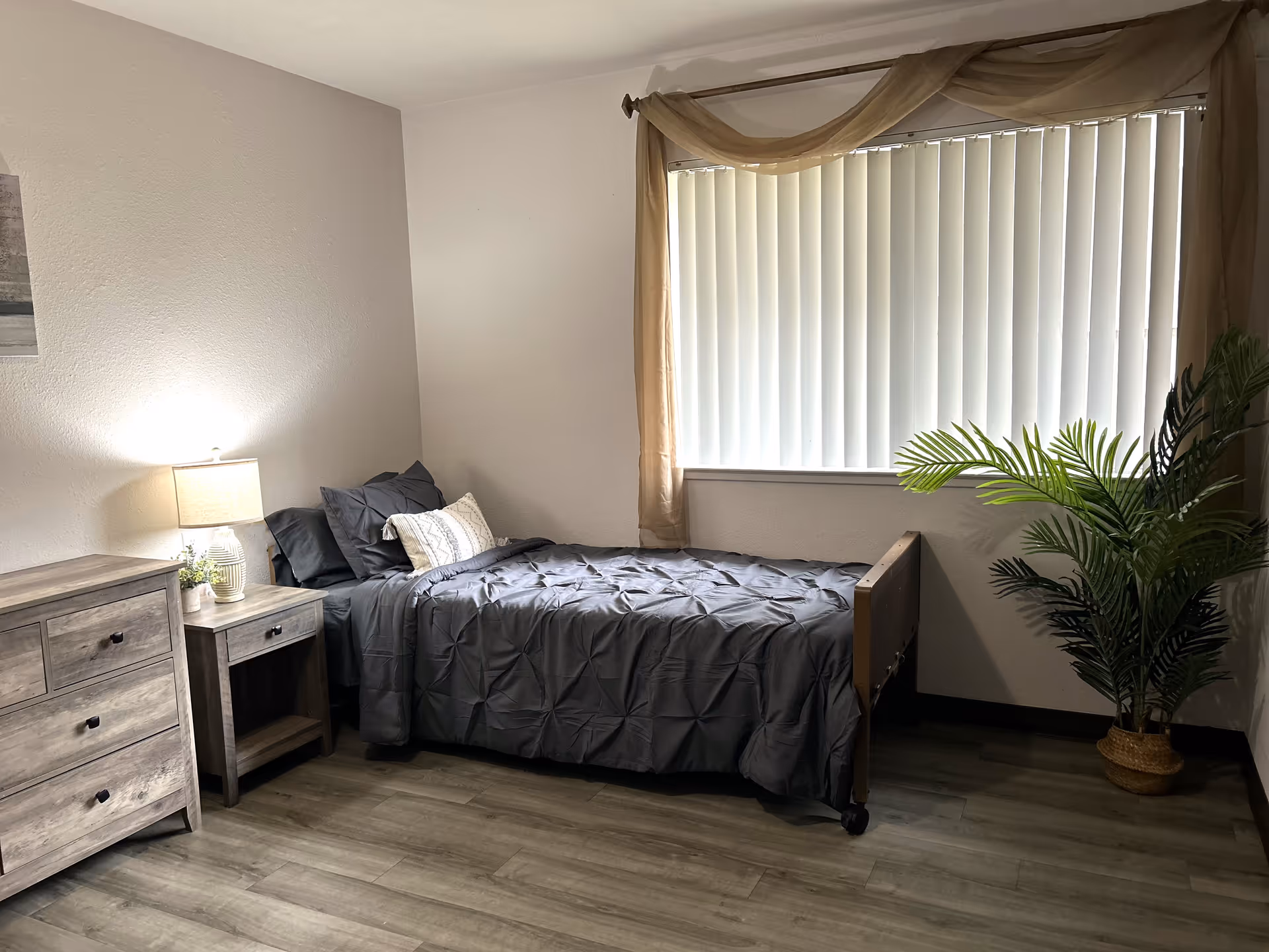 A simple bedroom with a single bed covered in a dark gray comforter and pillows. Next to the bed is a wooden nightstand with a lamp and a small plant. There is a wooden dresser on the left side of the image. A large window with vertical blinds and sheer brown curtains lets in natural light. A potted green plant is placed in the corner near the window. The floor is wood-style laminate.