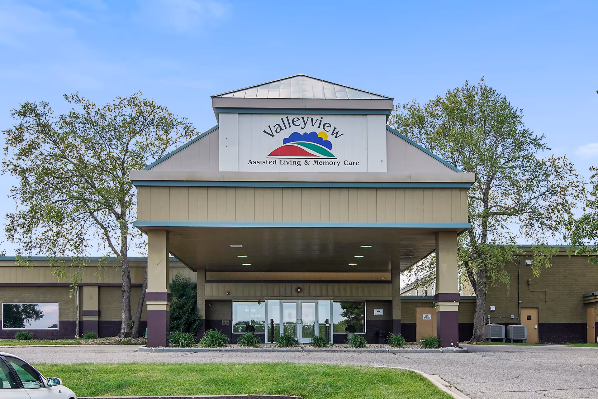 Front exterior view of the Valleyview Assisted Living & Memory Care building with a covered entrance, glass doors, and trees on either side under a clear blue sky.