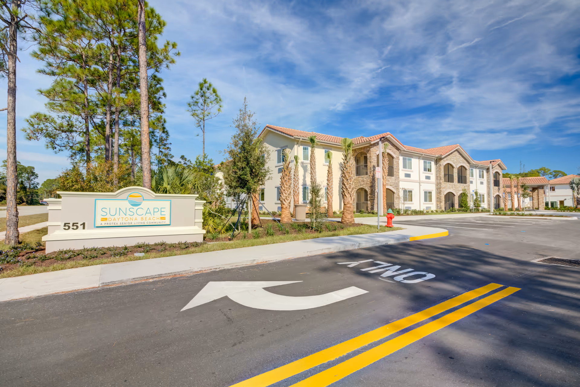 Front exterior of a two-story senior living community with a landscaped sign reading "SUNSCAPE Daytona Beach", palm trees, and a driveway with road markings.