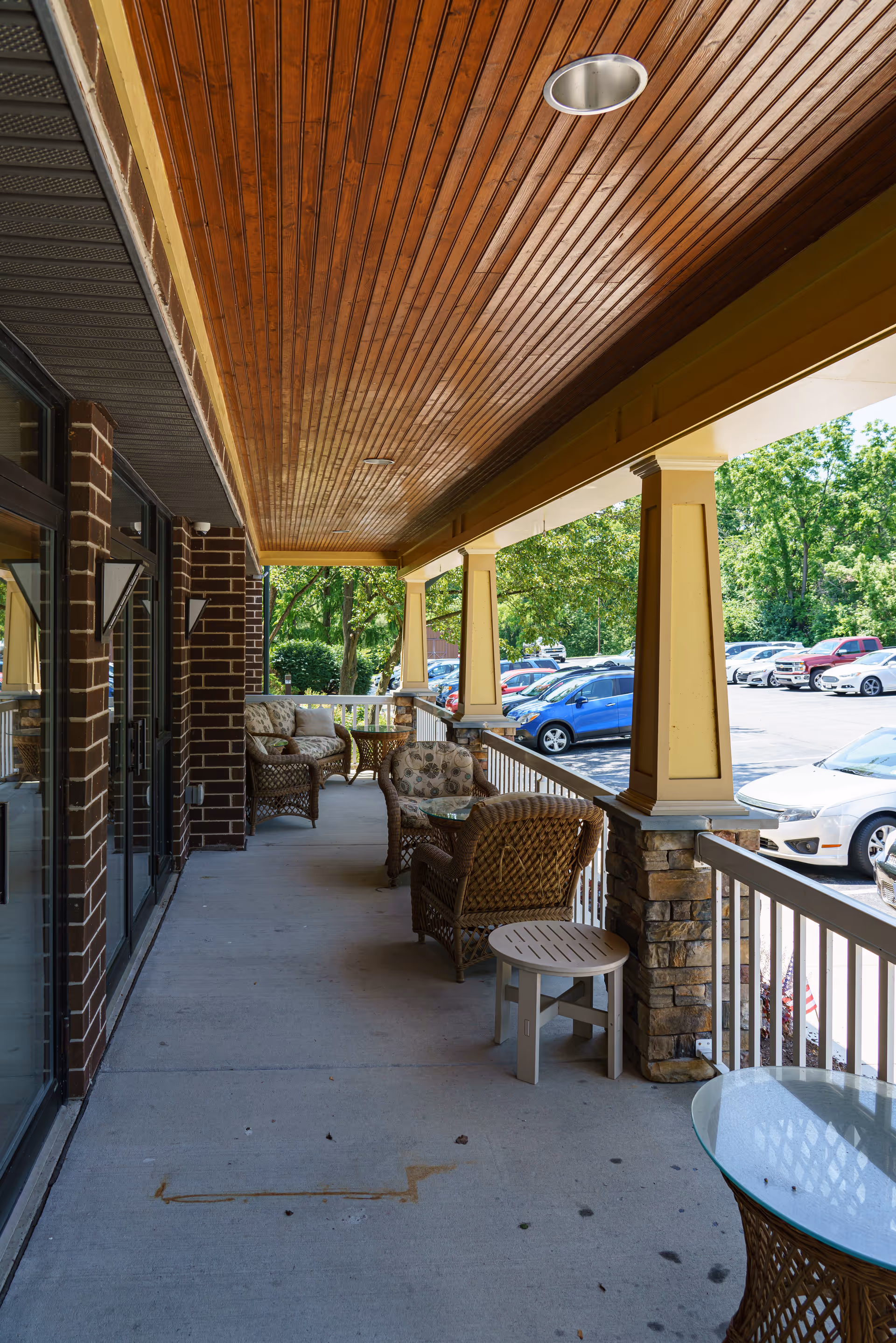 Covered front porch with wicker chairs and small tables overlooking a parking lot at a senior living facility.