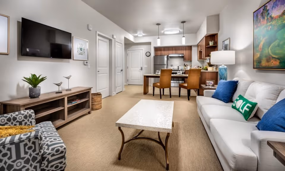 A modern living room and kitchen area in a senior living facility. The living room features a white sofa with blue and green pillows, a patterned armchair, a coffee table, and a wooden TV stand with a flat-screen TV mounted on the wall. The kitchen area includes a dining table with two brown chairs, stainless steel appliances, and wooden cabinets. The walls are decorated with framed artwork and a lamp on a side table next to the sofa.