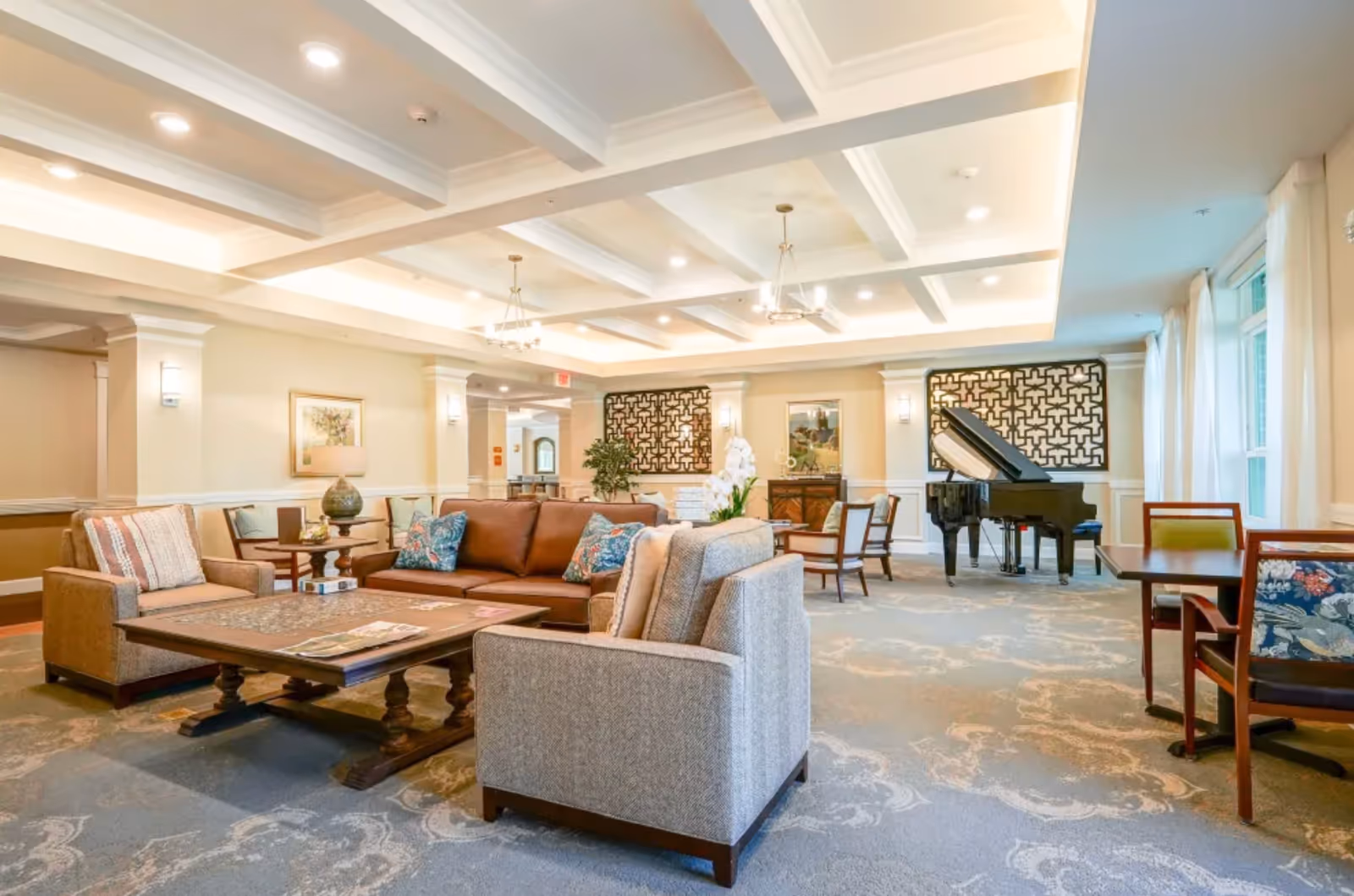 Spacious senior living community lounge with sofas, armchairs, a coffee table and a grand piano under a coffered ceiling.