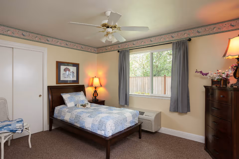 A small furnished bedroom with a twin bed covered in a blue quilt, nightstand with a lamp, dresser, window with curtains and a ceiling fan.