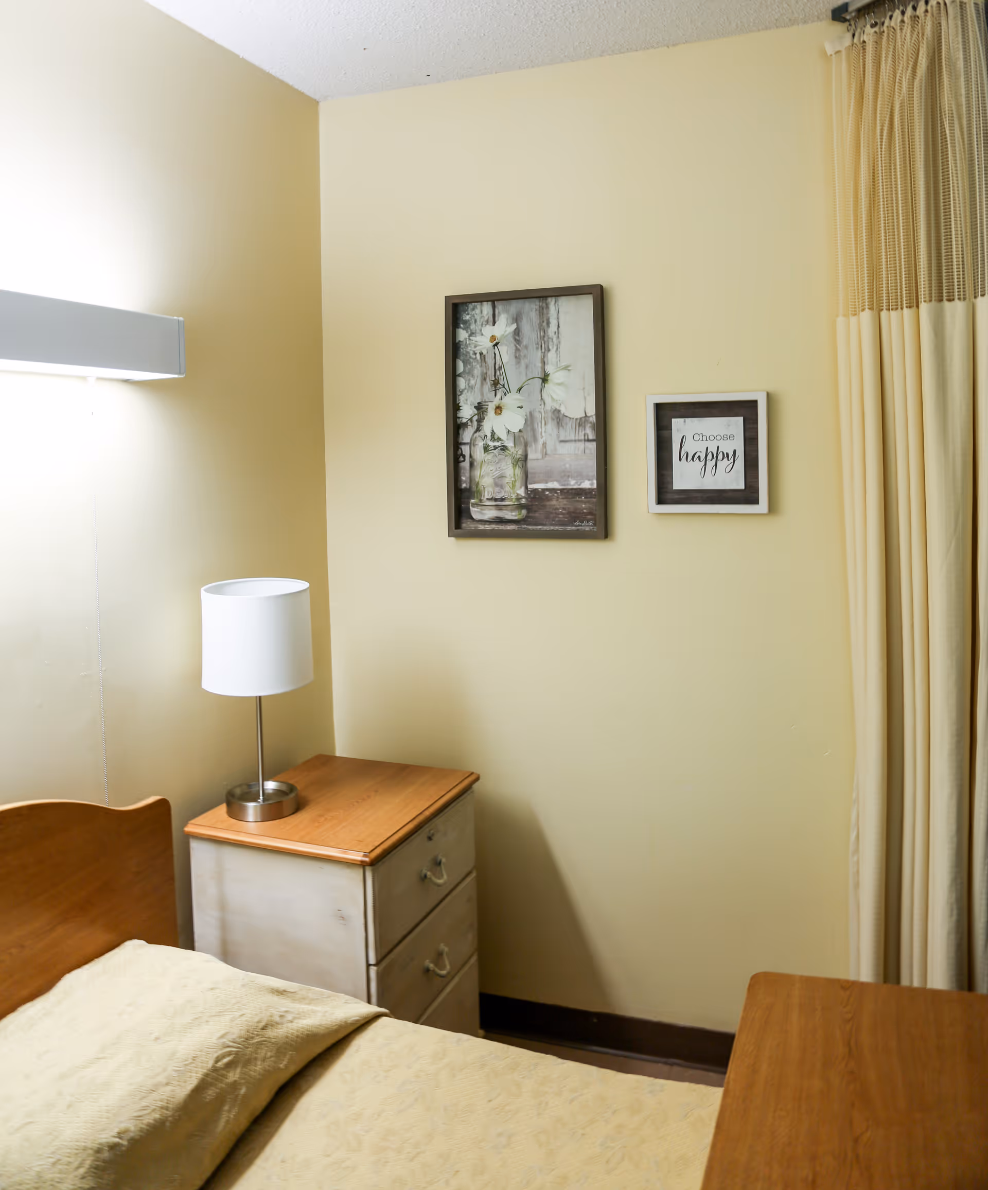 A small bedroom corner with a wooden bed partially visible, a nightstand with a lamp on top, two framed pictures on a pale yellow wall, and beige curtains on the right side.