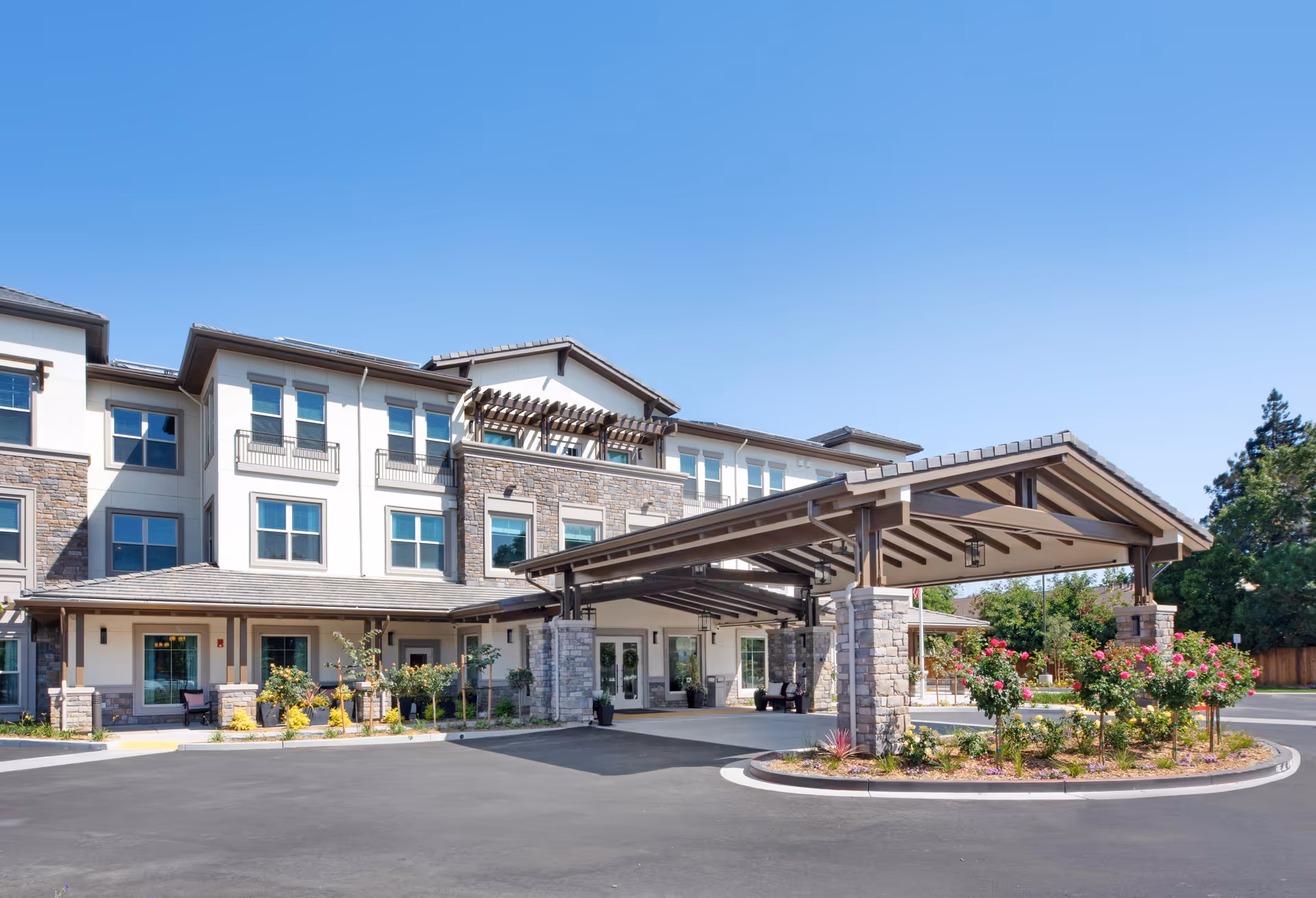 Three-story modern senior living facility entrance with a covered porte-cochere, stone accents, and landscaped driveway under a clear blue sky.