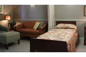A cozy room with a single bed covered in a floral bedspread, a brown sofa with cushions, a green armchair, a small lamp on a side table, and a potted plant in the corner. The walls are painted in neutral tones with white trim.