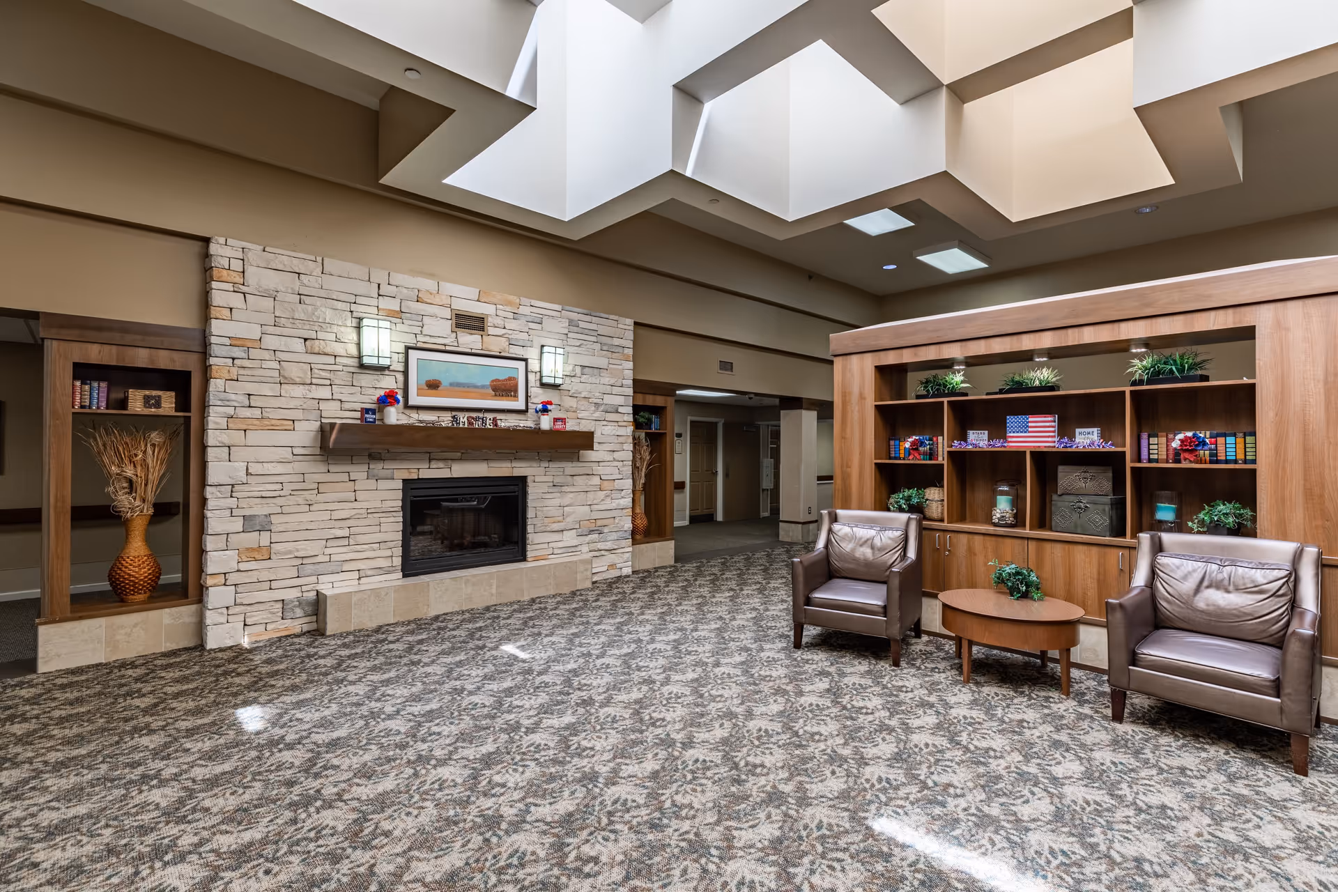 A cozy senior community lounge area featuring a stone fireplace with a wooden mantel, two brown leather armchairs, a small round wooden table with a plant, and a large wooden bookshelf decorated with plants, books, and patriotic-themed decor. The room has a patterned carpet and a ceiling with skylights allowing natural light to enter.