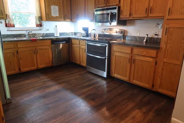 A kitchen with wooden cabinets, dark countertops, and stainless steel appliances including a dishwasher, oven, and microwave. There is a window with curtains above the sink, and the floor is dark wood.