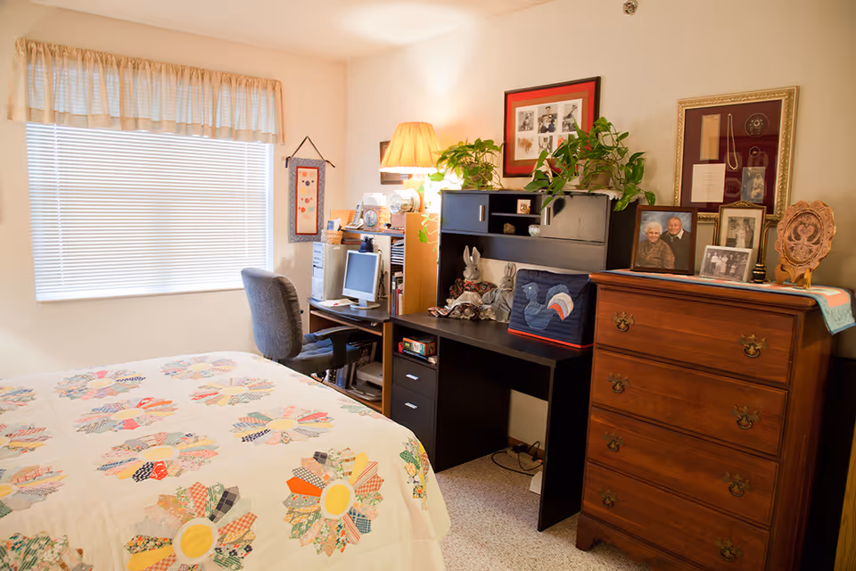 A cozy bedroom with a bed covered in a colorful floral quilt, a window with blinds and a valance, a desk with a computer and chair, a black hutch with plants and decorative items, and a wooden dresser with framed photos and artwork on the wall above it.
