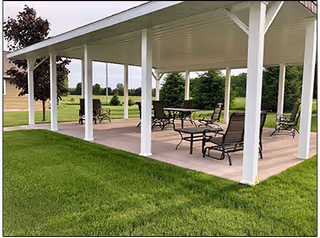 Outdoor covered patio area with multiple metal tables and chairs on a concrete floor, surrounded by green grass and trees in the background under a clear sky.
