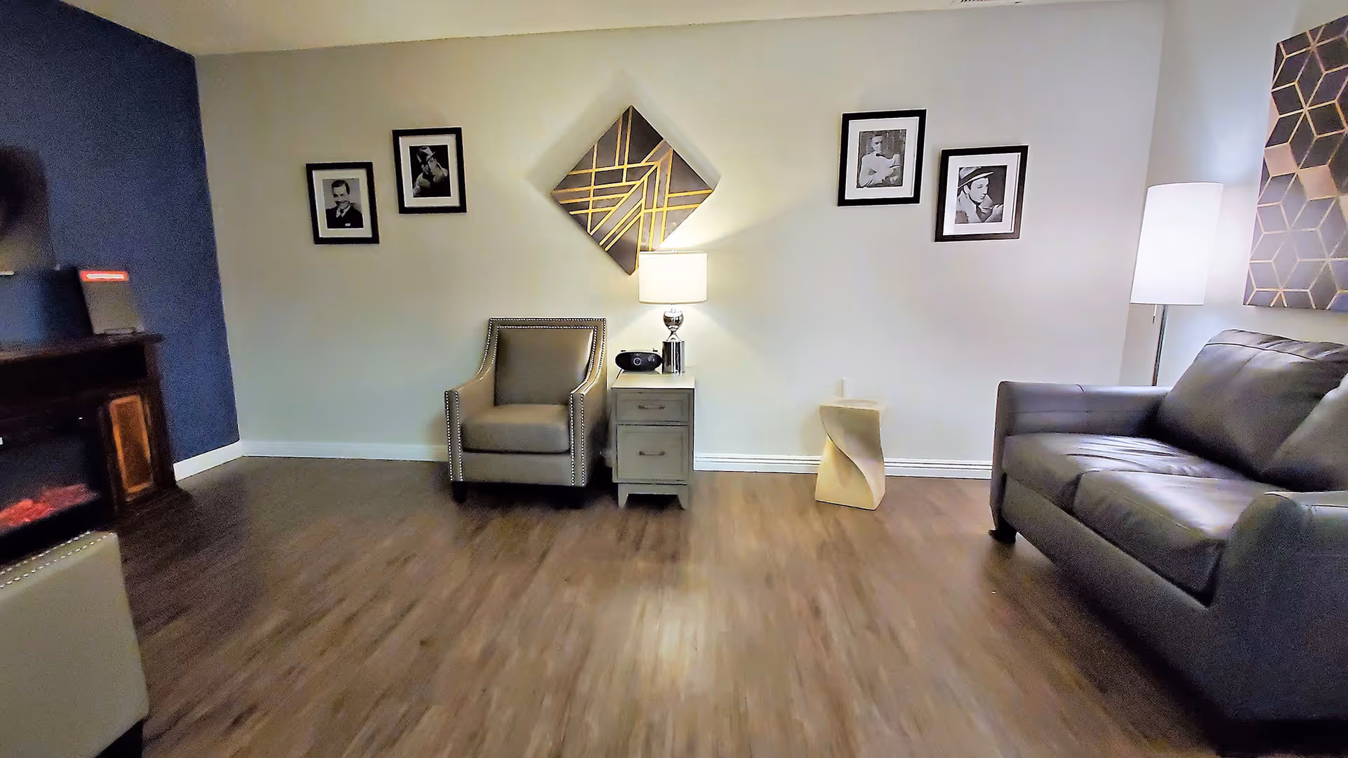 A cozy living room with wooden flooring, featuring a gray armchair and a gray sofa. Between them is a small side table with a lamp and a radio. The walls are decorated with framed black and white photos and a geometric art piece. A floor lamp stands next to the sofa, and a dark blue accent wall is visible on the left side.