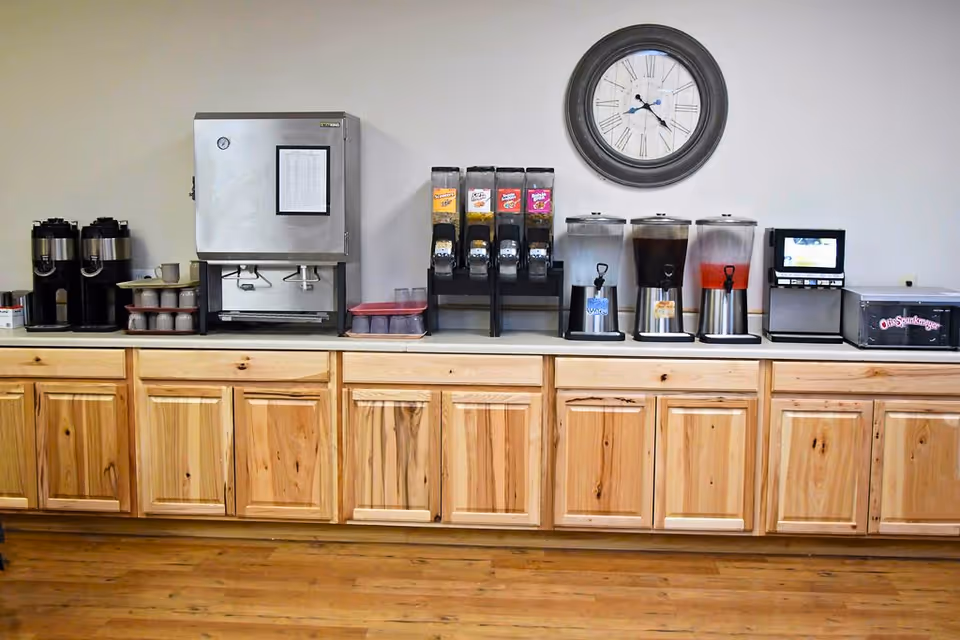A beverage station with wooden cabinets below and various drink dispensers on the countertop, including coffee urns, a hot water dispenser, cereal dispensers, juice dispensers, a small ice machine, and a wall clock above.