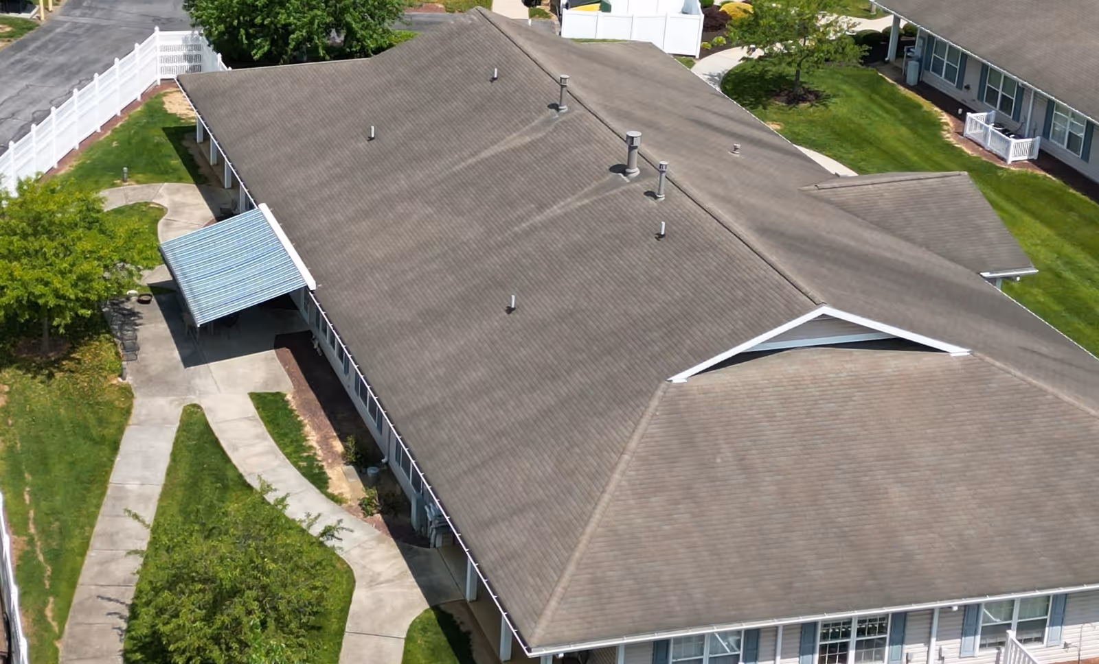 Aerial view of a senior living facility building with a large, brown shingled roof, surrounded by green lawns, trees, and paved walkways. There is a covered entrance on one side and white fencing along the perimeter.