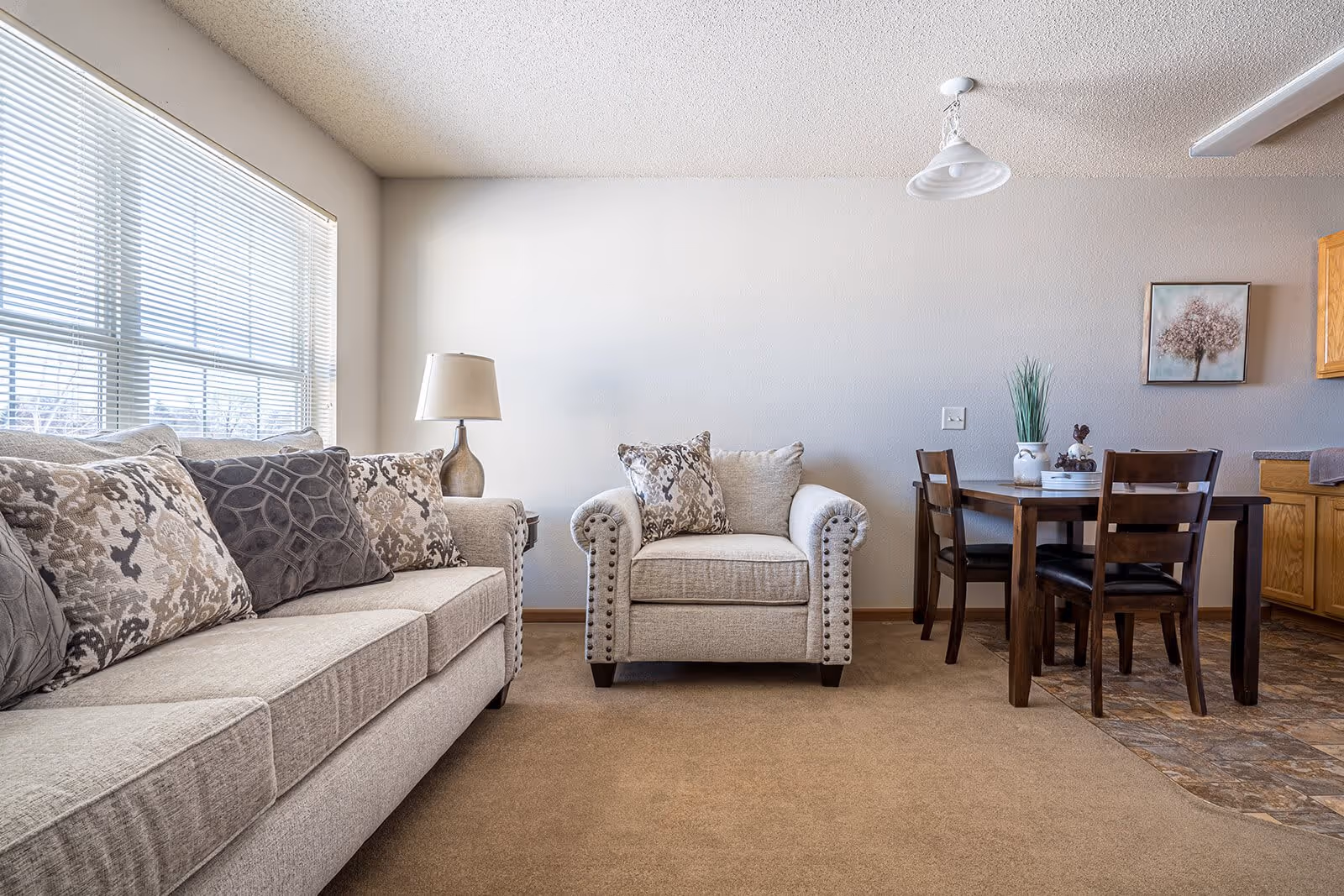Bright living room with a beige sofa and armchair by a large window, a lamp, and a small dining table with four chairs in the background.