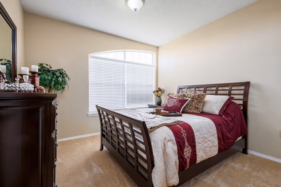 A bedroom with a wooden bed frame featuring a white and burgundy bedspread with decorative pillows. There is a large window with white blinds behind the bed. To the left, there is a dark wooden dresser with a mirror, candles, and a potted plant on top. The walls are beige and the floor is carpeted.