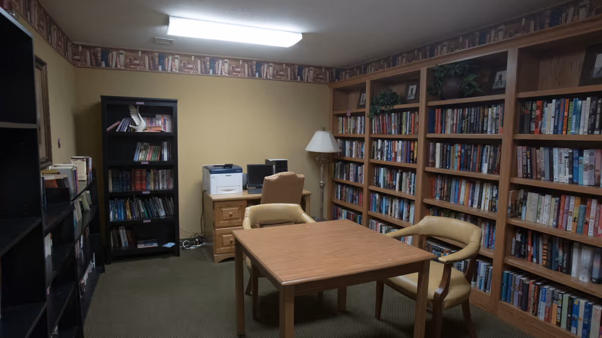 A small library or reading room with wooden bookshelves filled with books along two walls. There is a wooden table with two beige chairs in the center of the room. Against the back wall, there is a wooden desk with a computer, printer, and a beige office chair. A floor lamp stands next to the desk. The walls are painted yellow with a wallpaper border featuring books near the ceiling.