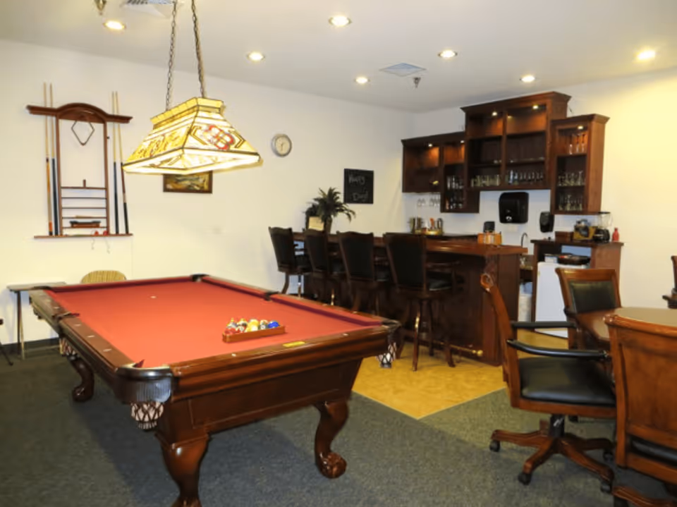A recreational room featuring a red felt pool table with a rack of billiard balls, a hanging stained glass light fixture above the table, a wooden bar with four bar stools, and a wall-mounted rack holding pool cues. There are also several chairs and a small table in the foreground, with shelves behind the bar holding glassware and other items.