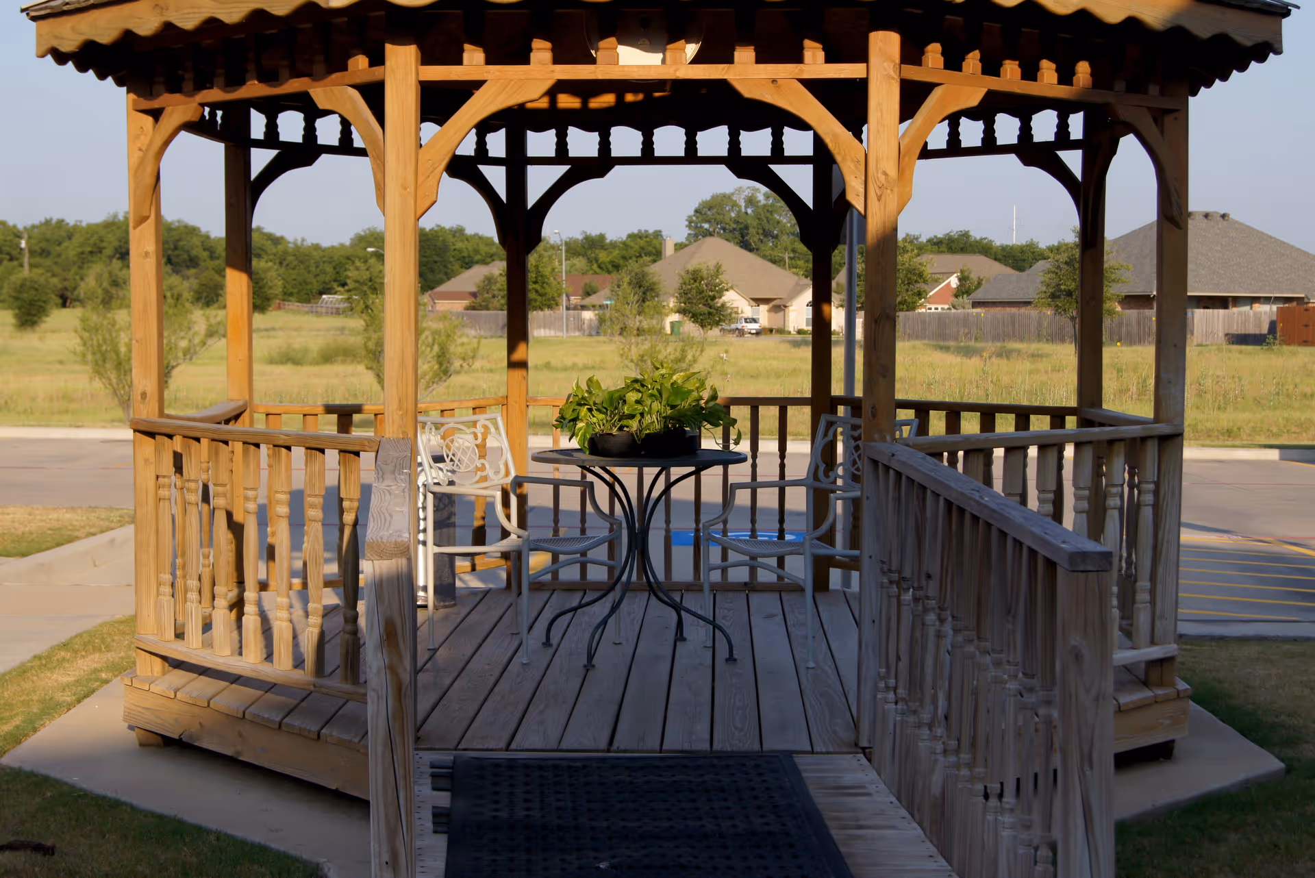 A wooden gazebo with a small round table and two white metal chairs inside. A potted green plant is on the table. The gazebo is situated outdoors with a grassy field and houses visible in the background.