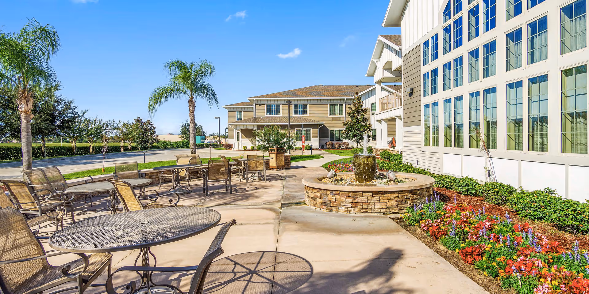 Outdoor patio courtyard with multiple tables and chairs, a central stone fountain, landscaped flowerbeds, palm trees, and the adjacent building with large windows.