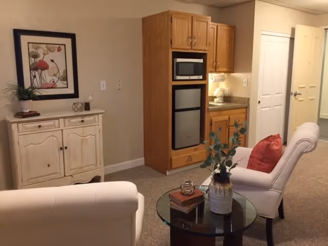 A cozy interior living space featuring two upholstered armchairs with a red pillow on one, a round glass coffee table with a decorative vase and books, a wooden cabinet with a potted plant and framed artwork above it, and a built-in wooden kitchenette area with a microwave, mini refrigerator, and countertop with a small flower arrangement. A hallway with doors is visible in the background.