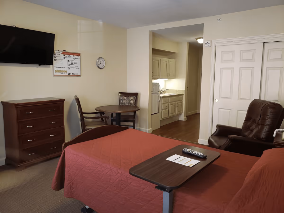 Studio-style senior living bedroom with a red-covered bed, overbed table, wall-mounted TV, dresser, small dining table, recliner and a kitchenette in the background.