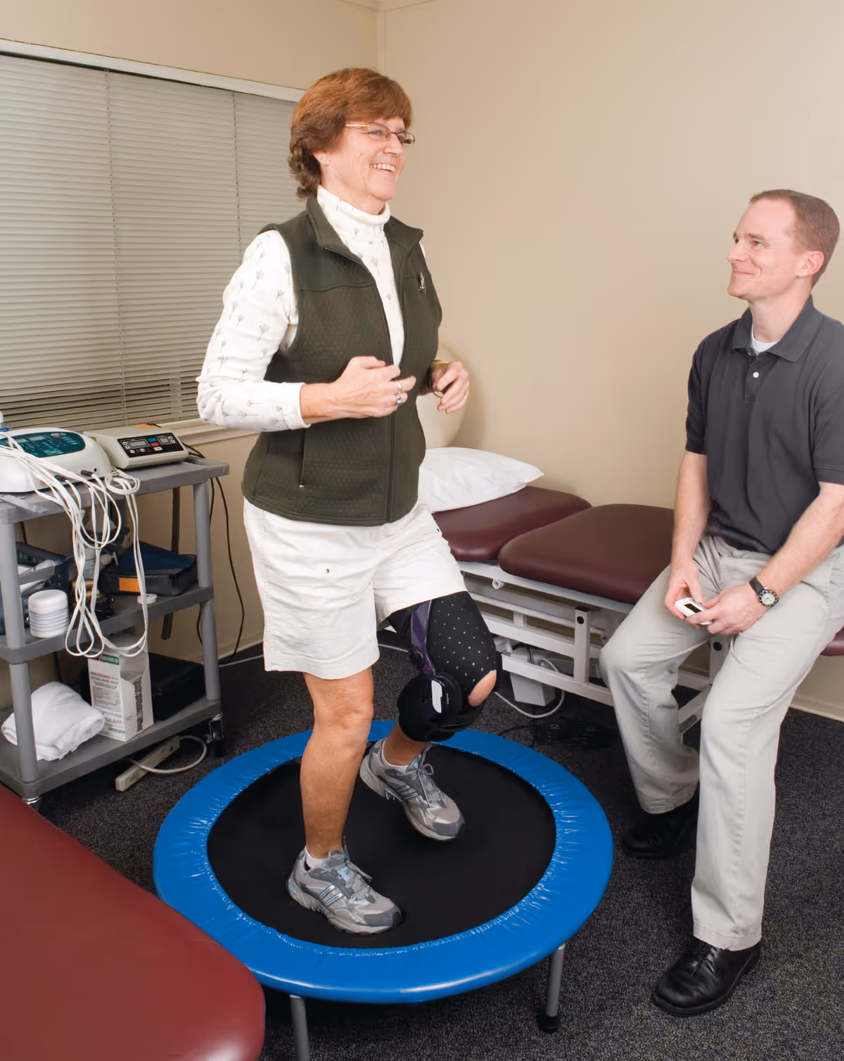 An elderly woman wearing a knee brace is exercising on a small trampoline in a medical or rehabilitation room while a man, possibly a therapist, watches and smiles. The room contains medical equipment and examination tables.