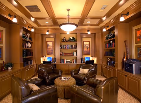 A cozy interior room with four brown leather armchairs arranged around a round ottoman. The room features built-in wooden bookshelves filled with books and decorative items, two computer desks with monitors, warm lighting from wall sconces and a ceiling light, and framed artwork on the walls.