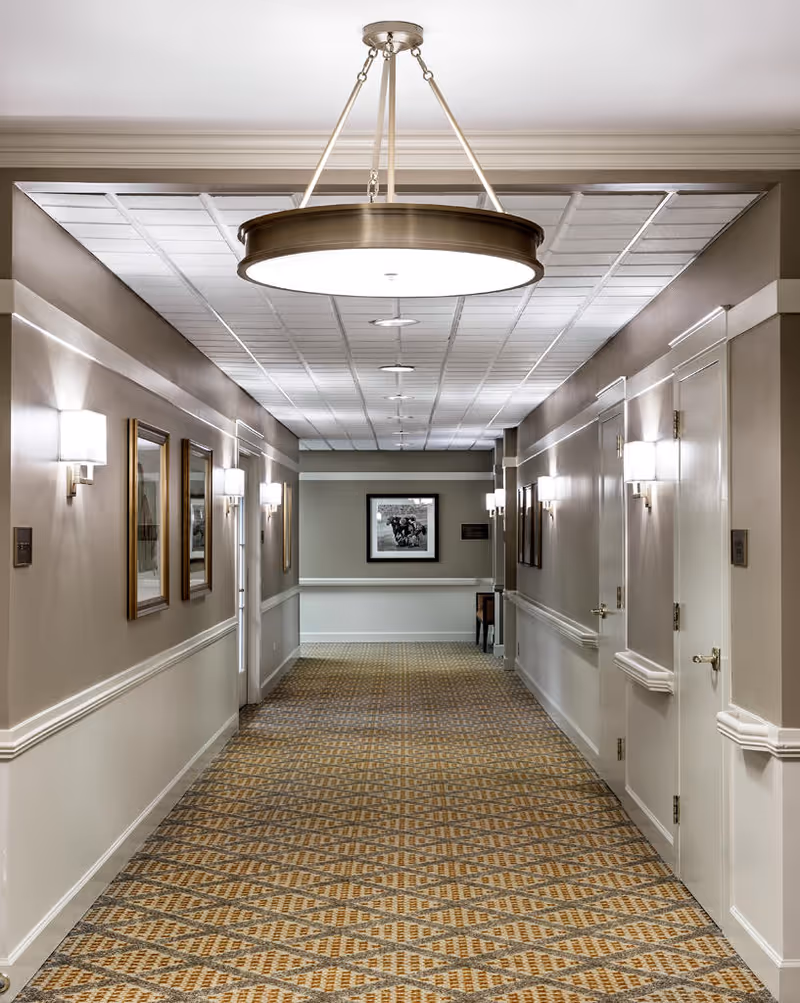 A well-lit interior hallway with patterned carpet, beige walls with white trim, wall-mounted lights, framed pictures on the walls, and a large circular ceiling light fixture. Several closed doors line the hallway.