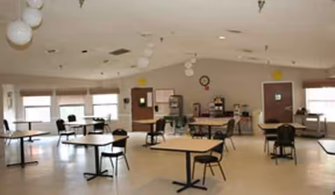 Empty senior living dining area with scattered tables, chairs, and large windows.