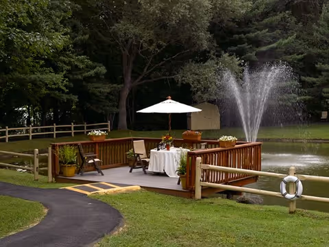 Wooden deck with a table set under an umbrella, chairs and potted plants beside a pond with a fountain and trees.