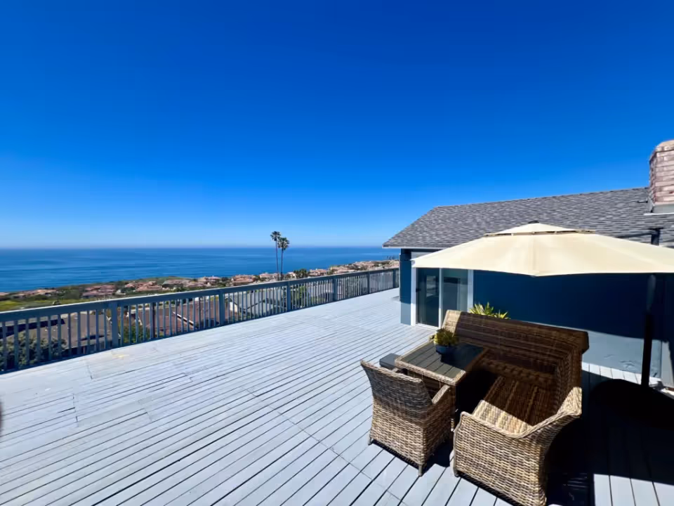 Spacious outdoor deck with wicker patio furniture including a table, two chairs, and a bench under a large beige umbrella, overlooking a scenic ocean view with clear blue sky and distant houses.
