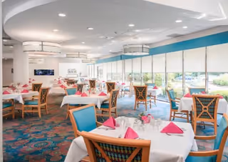Bright dining room with multiple tables covered in white tablecloths and pink napkins, wooden chairs with blue cushions, large windows with blinds letting in natural light, and modern ceiling lights.