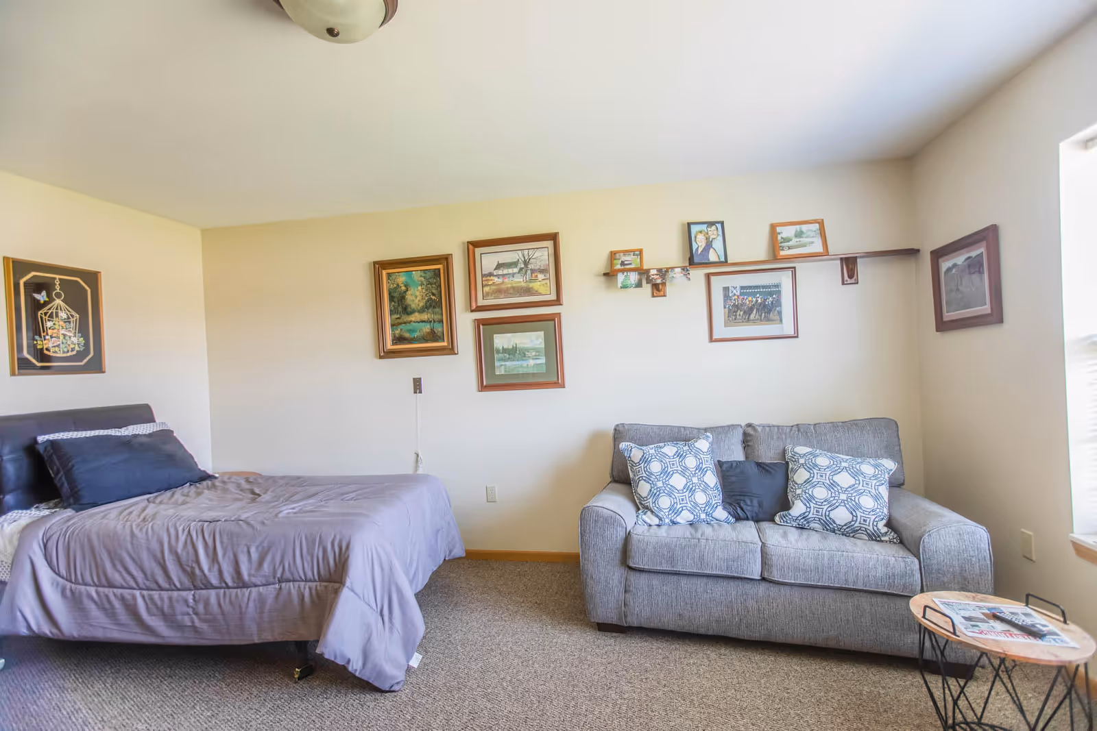 A cozy bedroom with a bed on the left side covered with a gray comforter and black pillows. On the right side, there is a gray loveseat with three decorative pillows. The walls are adorned with several framed pictures and artwork. A small round side table with a magazine is placed near the loveseat. The room has beige walls and carpeted flooring, with natural light coming in from a window on the right.