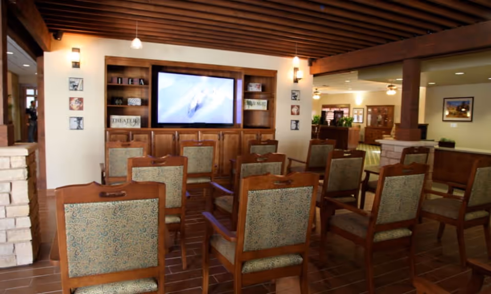 A small theater-style common room with rows of wooden chairs facing a large wall-mounted television and built-in shelving.