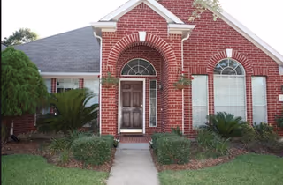 Front exterior of a red brick single-story house with an arched entryway, wooden door, flanking windows, and landscaped lawn.