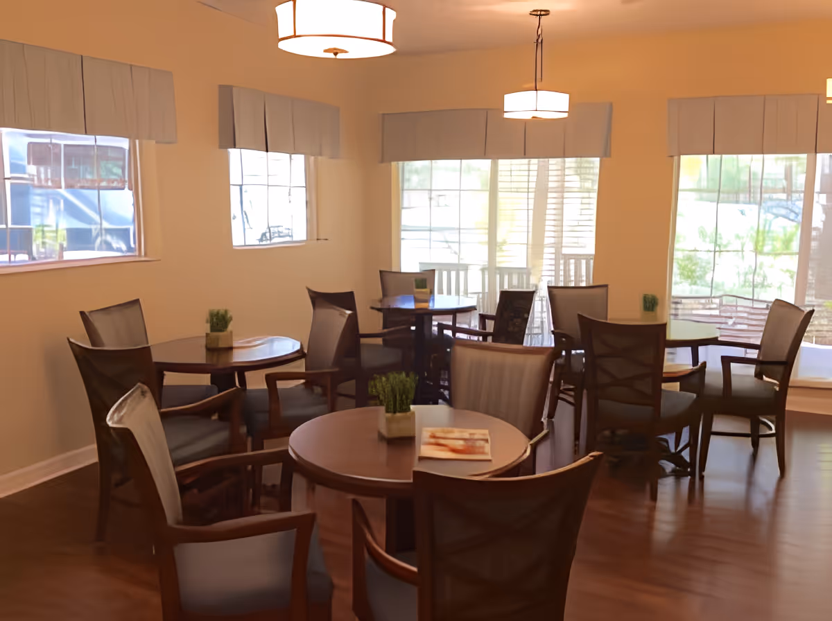 A bright dining room with several round wooden tables and cushioned chairs arranged around them. Each table has a small decorative plant and some have menus or books. Large windows with white blinds allow natural light to fill the room, and two ceiling light fixtures hang above the tables. The floor is wooden, and the walls are painted a light color.
