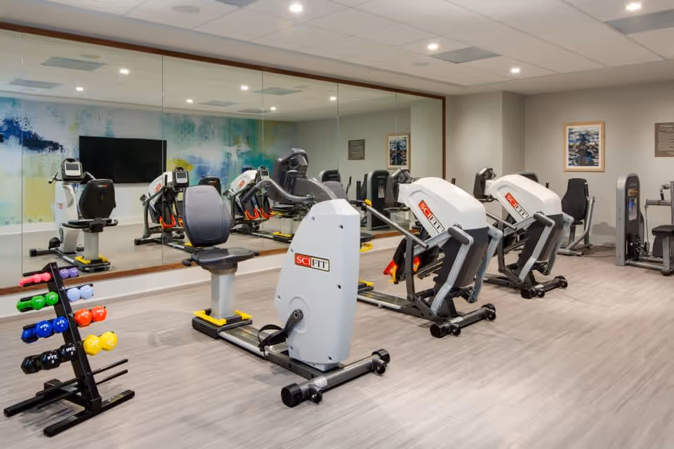 A bright fitness room with recumbent exercise bikes, a rack of colorful dumbbells, a large mirror and a wall-mounted TV.