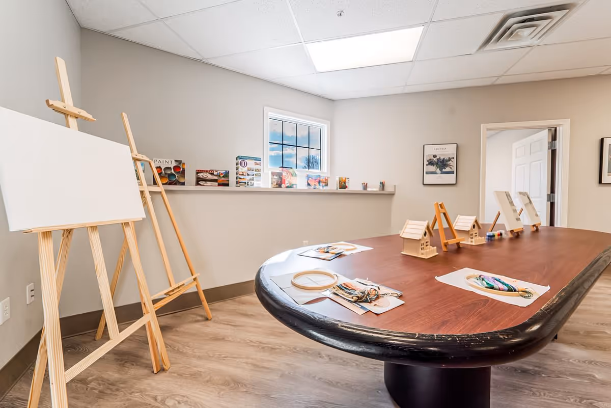 A bright art room with a large wooden table set up with small easels, wooden birdhouse models, embroidery hoops, and colorful yarn. Two blank wooden easels stand against the wall, and a shelf below a window holds various art supplies and kits. The room has light-colored walls and wood flooring.
