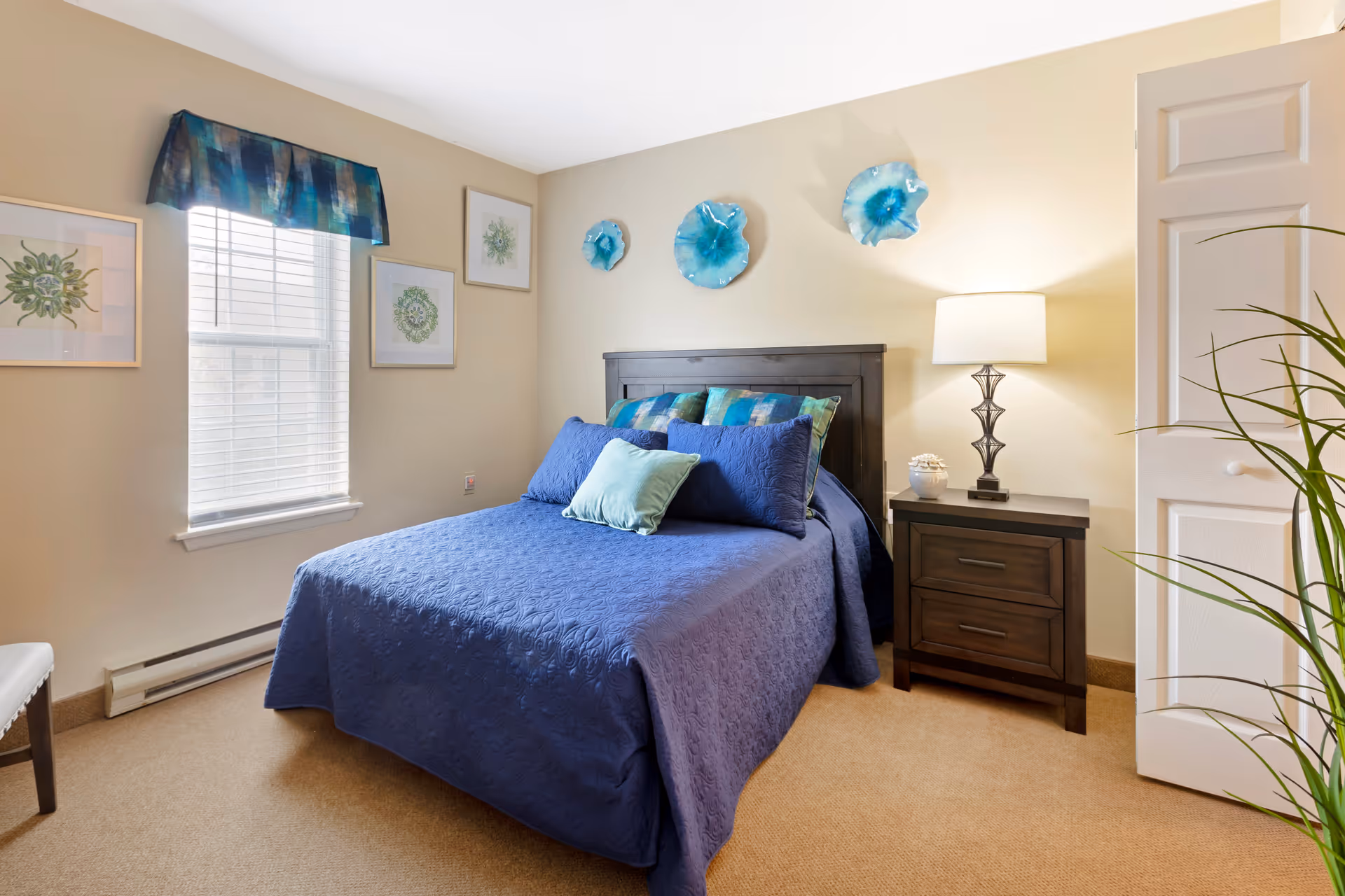 A cozy bedroom with a bed covered in a blue quilt and multiple pillows. There is a dark wooden headboard and matching nightstand with a lamp and decorative items. The walls are beige with three blue decorative wall hangings above the bed and framed artwork on the adjacent wall. A window with blinds and a blue valance lets in natural light. A plant is partially visible on the right side of the image.