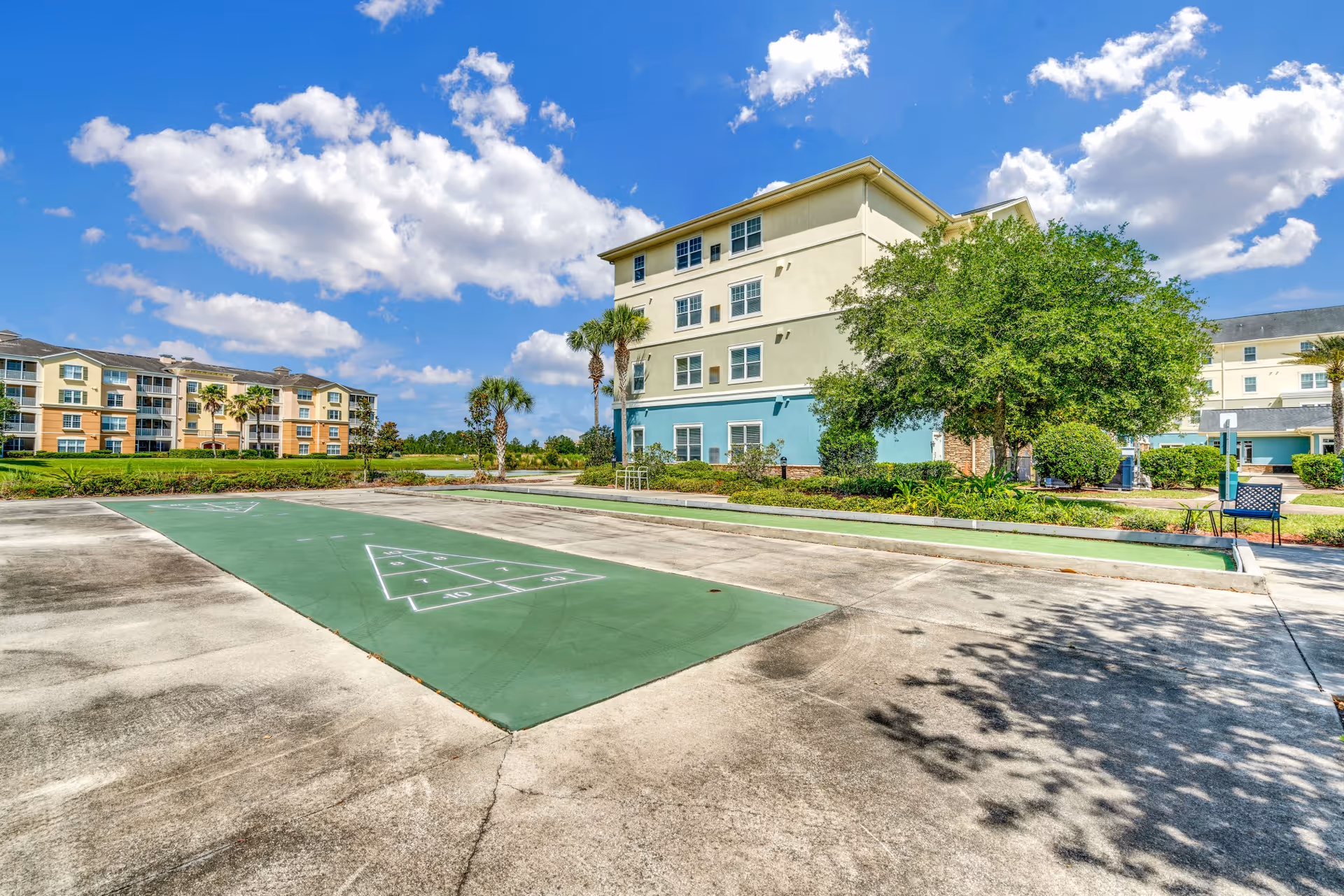 Outdoor shuffleboard courts with green playing surfaces and white markings, surrounded by concrete walkways, palm trees, and shrubbery. In the background, there are multi-story residential buildings under a blue sky with scattered clouds.