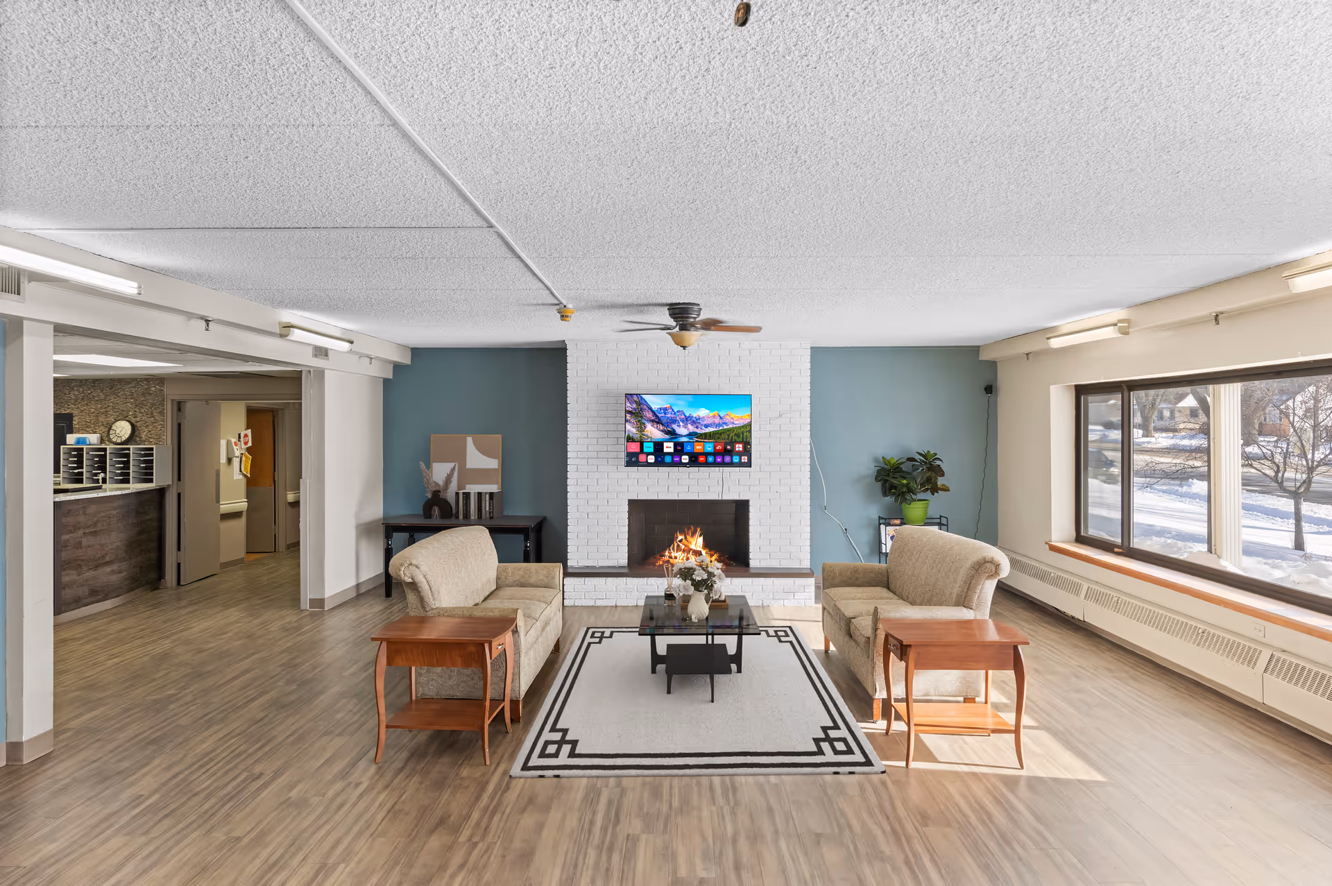 A bright and spacious living room area in a senior living facility with two beige sofas facing each other across a glass coffee table on a patterned rug. A white brick fireplace with a flat-screen TV mounted above it is centered on a teal accent wall. There are wooden side tables next to each sofa, a potted plant on a small stand to the right, and large windows on the right side letting in natural light. The floor is wood laminate, and the ceiling has a textured finish with fluorescent lighting.