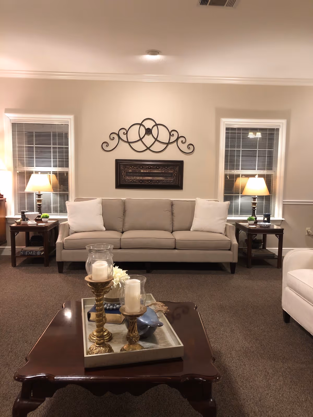 A cozy living room with a beige sofa adorned with two white pillows, flanked by two wooden side tables each holding a lamp. Above the sofa is decorative wall art featuring a metal scroll design and a framed piece. In front of the sofa is a dark wooden coffee table with a tray holding two candle holders with white candles and a small decorative item. The room has beige walls, two windows with blinds, and carpeted flooring.