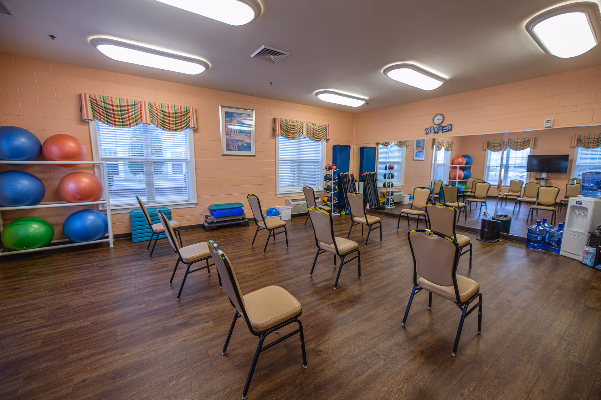 A spacious exercise room with wooden flooring, several beige chairs arranged in a circle, large mirrors on one wall, exercise balls on shelves, and various fitness equipment. The room has peach-colored walls, windows with striped valances, and ceiling lights.