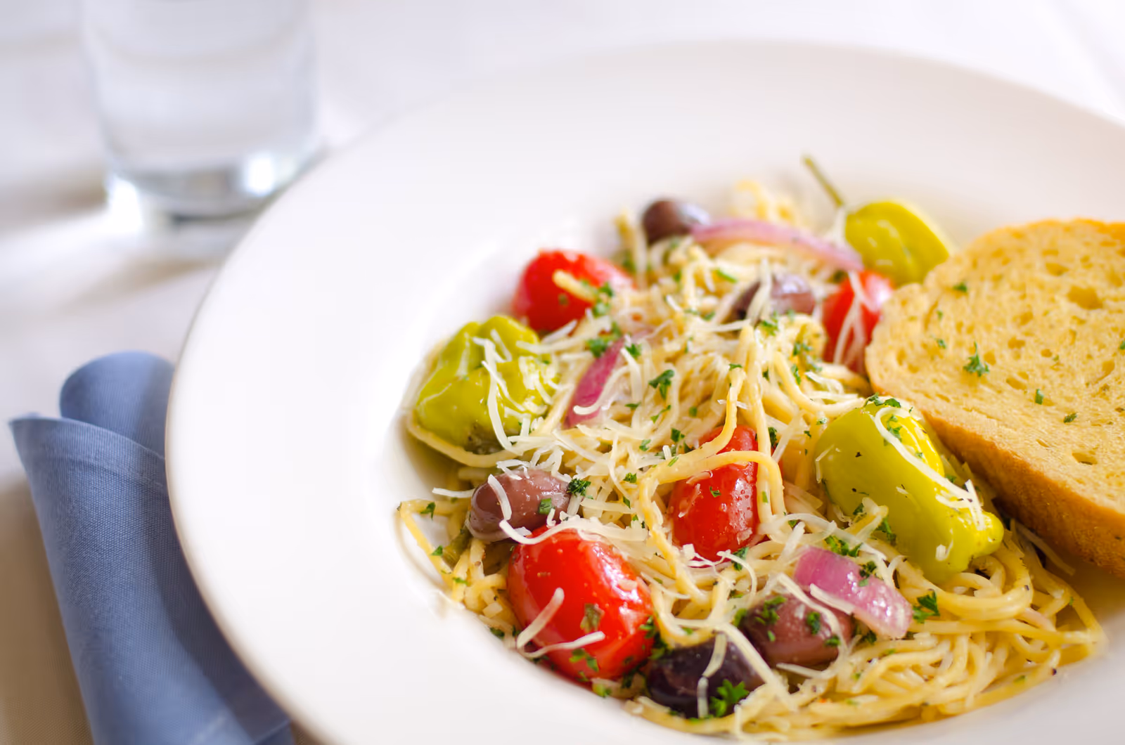 Close-up of a pasta salad with cherry tomatoes, peppers, olives, red onion, grated cheese and a slice of bread on a white plate.