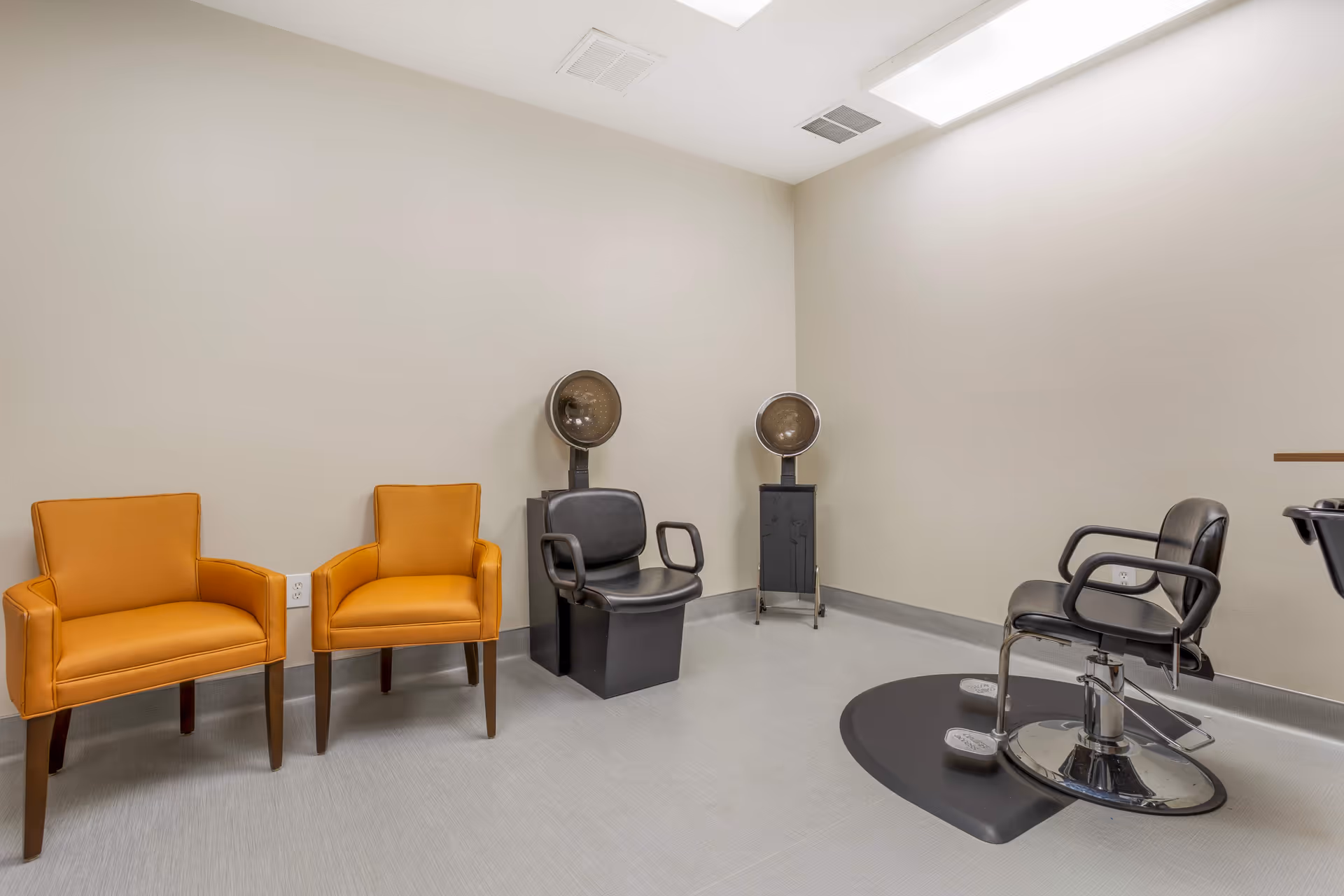 A small salon room with two orange chairs, two black hair drying chairs with hooded dryers, and a black salon styling chair on a gray floor with beige walls.