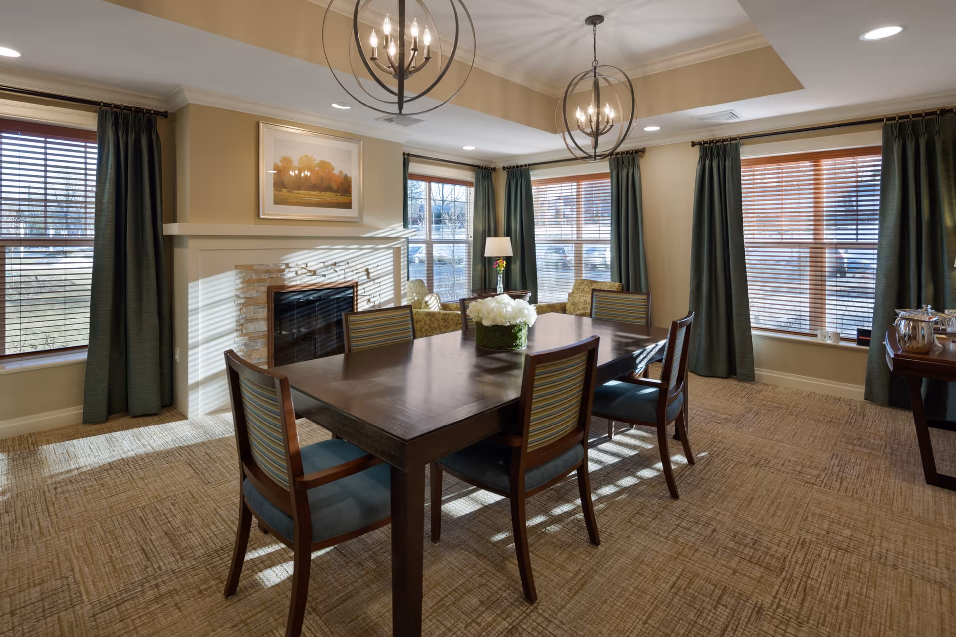 A well-lit dining room with a large wooden table surrounded by six chairs with striped upholstery. The room features large windows with wooden blinds and dark green curtains, a fireplace with a framed landscape painting above it, two modern spherical chandeliers, and a side table with a silver tea set. The carpet is beige with a subtle pattern.